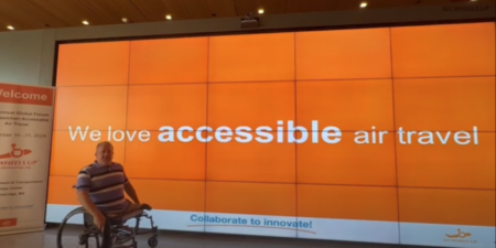 Dave Stevens, a board member of All Wheels Up, in front of a giant screen at the Global Forum, showing the words 'We love accessible air travel' against an orange background